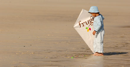 A toddler on a beach holding a kite featuring the frugi brand.