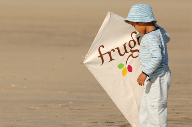 A toddler on a beach holding a kite featuring the frugi brand.
